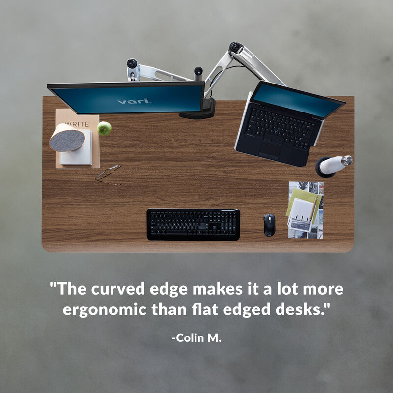 Overhead view of a walnut desktop with a monitor and laptop on an arm, keyboard, mouse and books. Customer review text reads: &ldquo;The curved edge makes it a lot more ergonomic than flat edged desks.&rdquo; Colin M. image number null